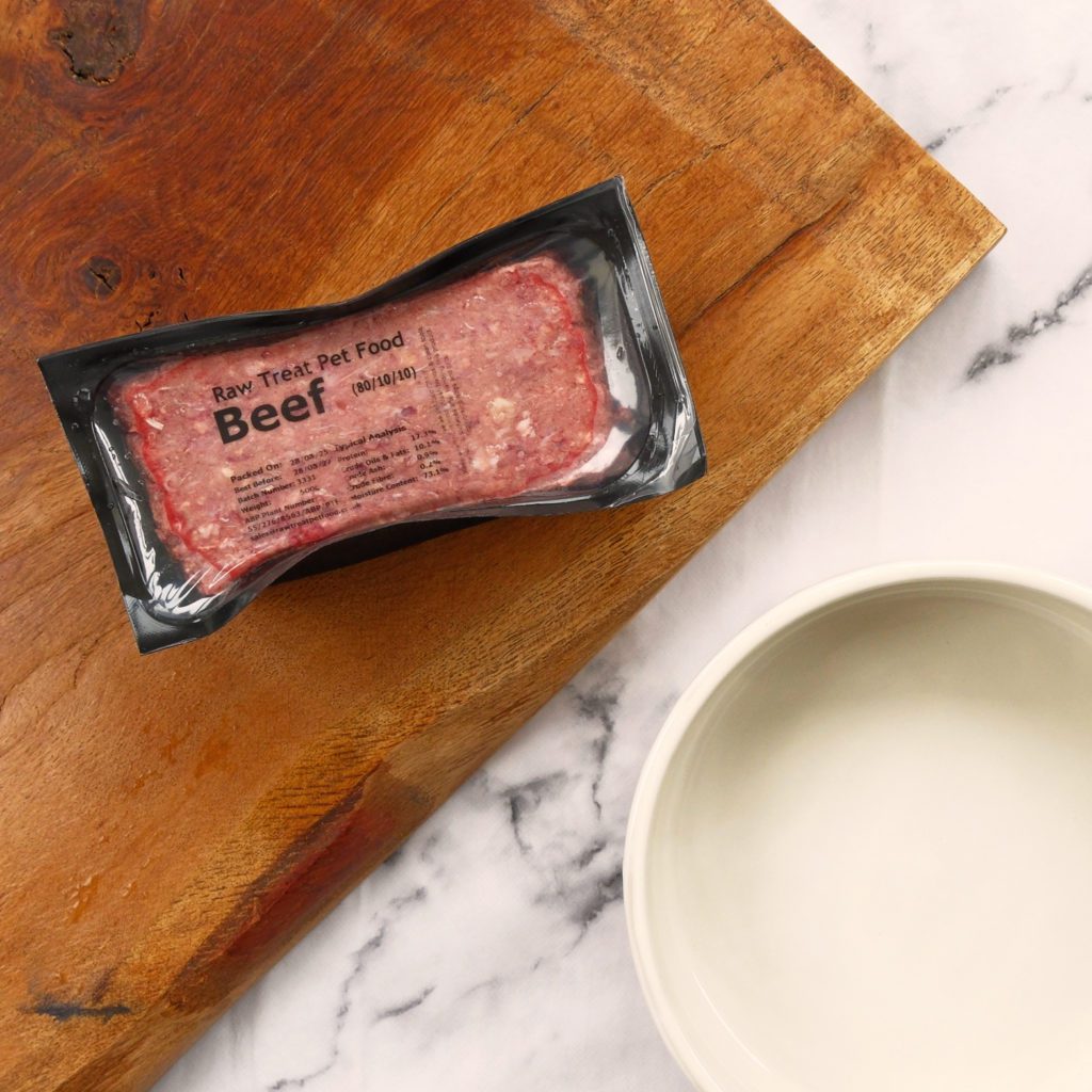 A pack of Raw Treat Pet Food sat on a wooden board next to a dog food bowl