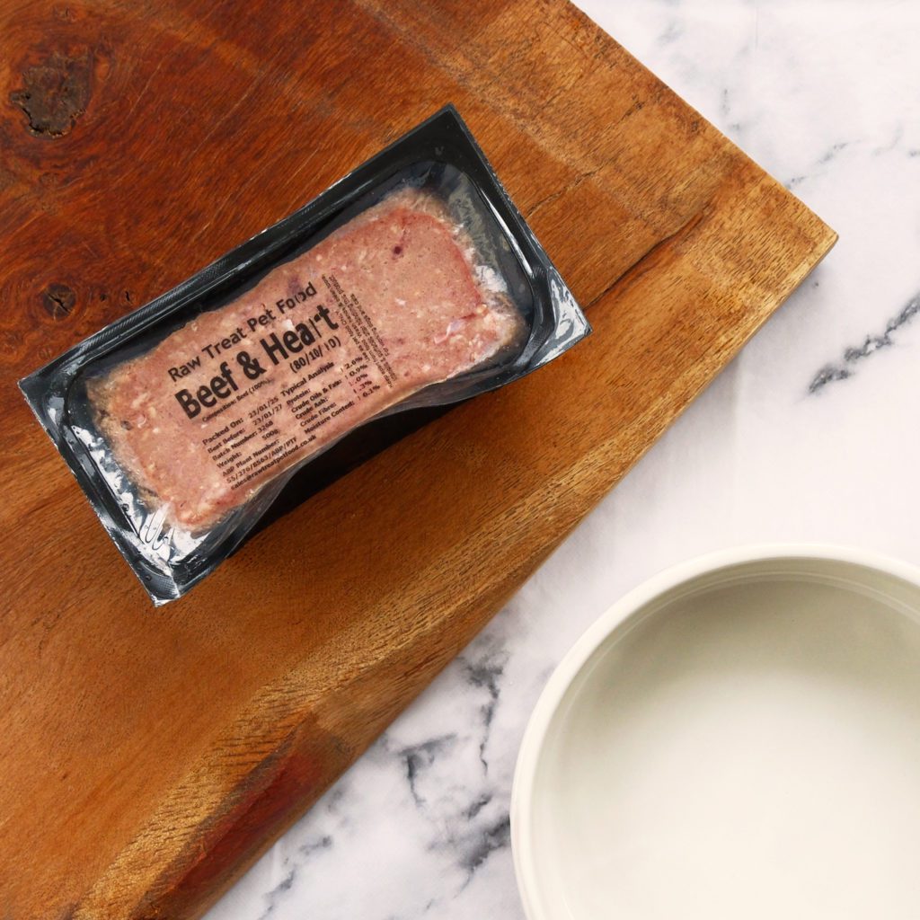 A pack of Raw Treat Pet Food sat on a wooden board next to a dog food bowl