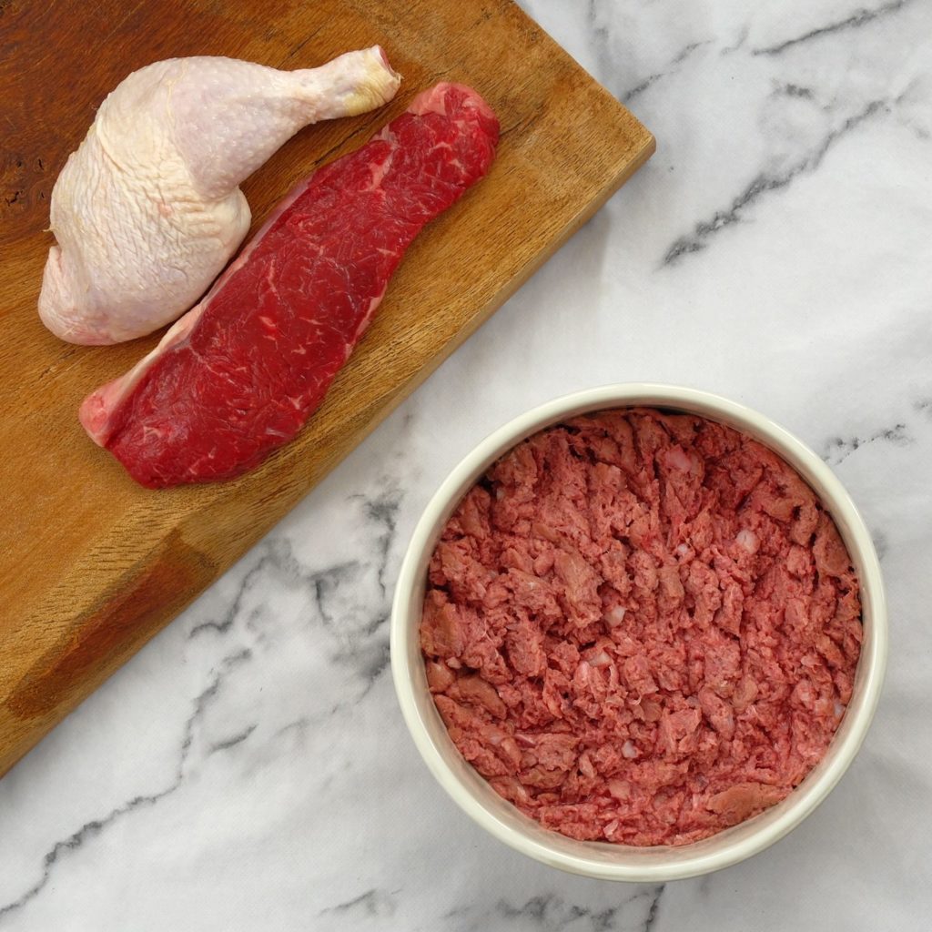 Beef and chicken mince in a dog bowl with a chicken leg and beef steak on a chopping board