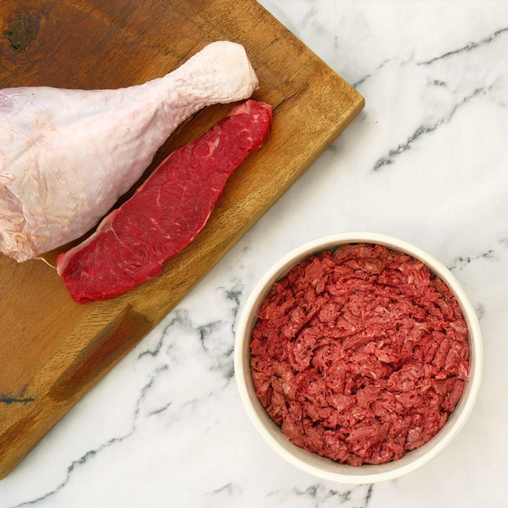Beef and turkey mince in a dog bowl with a beef steak and turkey leg on a chopping board